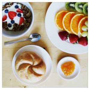 Eine Schüssel mit Müsli, ein Teller mit Früchten, eine Semmel und eine Portion Marmelade befinden sich auf einem Tisch.