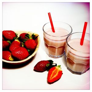 Links ist eine kleine Schüssel mit Erdbeeren, rechts zwei Erdbeer-Yoghurtdrinks mit roten Strohhalmen.