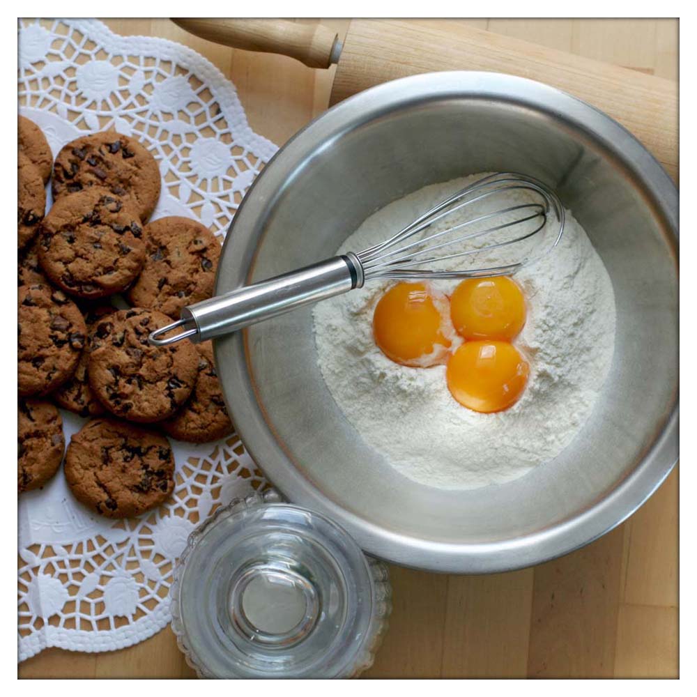 Links im Bild sind fertige Kekse mit Schokoladestückchen, rechts im Bild eine Metallschüssel in welcher Mehl, drei Eigelb und ein Schneebesen.