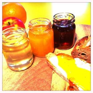 Drei Gläser mit Konfitüre und daneben ein Laib Brot. Links hinten liegt ein Apfel und eine Orange.