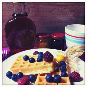 Waffeln mit Waldbeeren auf einem Teller, im Hintergrund eine Flasche Ahornsirup und zwei kleine Gläser Marmelade.