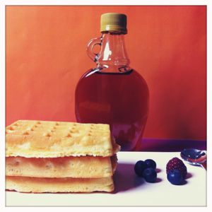 Auf einem Teller liegen drei Waffeln, daneben ein paar Waldbeeren. Im Hintergrund steht eine Flasche Sirup.